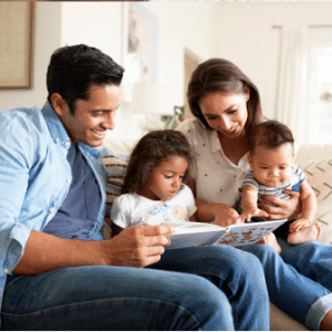 A smiling family sitting on a couch, reading a book together, with two young children.