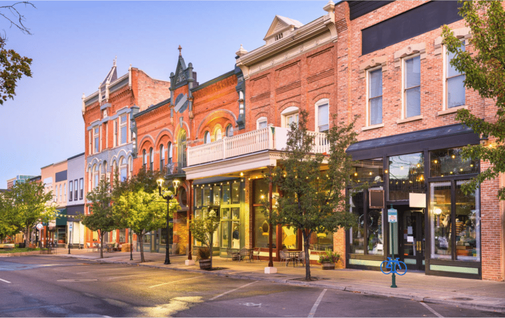 A row of historic brick storefronts in a small town, illuminated by streetlights during early evening, with trees lining the sidewalk.