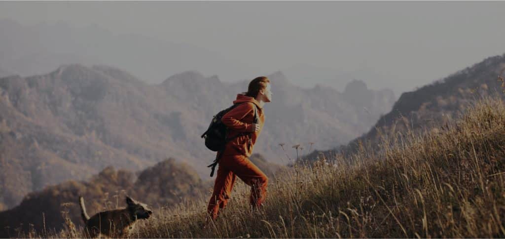 A person hiking uphill on a grassy slope with a backpack, followed by a dog, with mountains in the background under soft daylight.